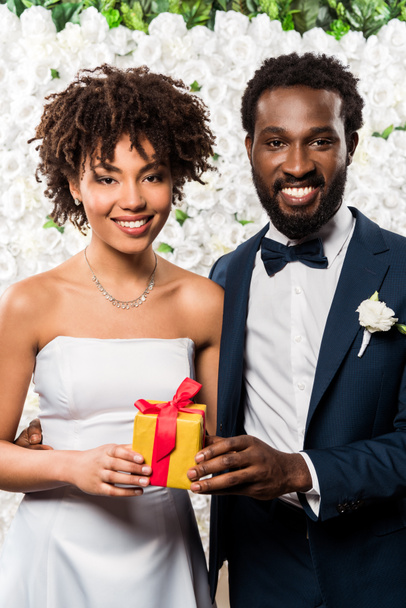 happy african american bride holding present near bridegroom and flowers  - Fotografie, Obrázek