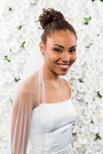happy african american bride in white veil smiling near flowers  - Foto, Bild