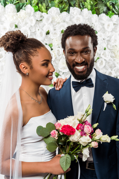 happy african american bride in white veil smiling near bridegroom and flowers  - Photo, Image