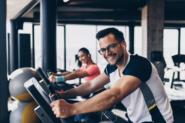 Young and attractive woman and man biking in the fitness gym. They exercising legs and doing cardio workout while riding cycling machines. - Fotó, kép