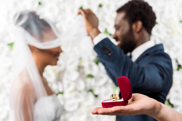 アフリカ系アメリカ人のカップルの近くに結婚指輪を持つ箱を持つ男のトリミングされたビュー  - 写真・画像