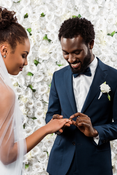 l'uomo afro-americano allegro che mette l'anello nuziale al dito di sposa vicino a fiori
  - Foto, immagini