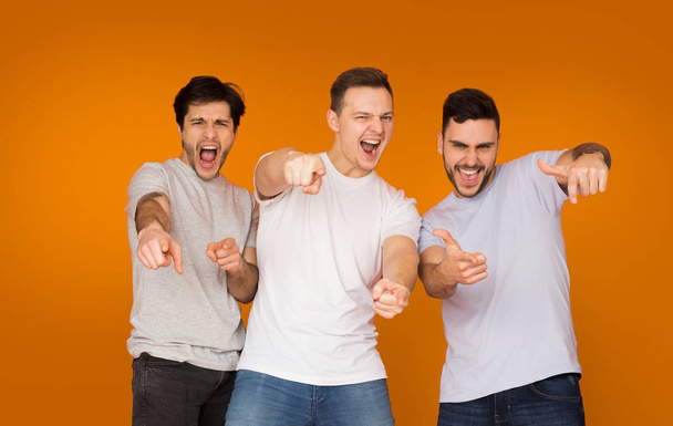 Ενθουσιασμένοι φίλοι δείχνοντας τα δάχτυλά σας στην κάμερα, πορτοκαλί φόντο - Φωτογραφία, εικόνα