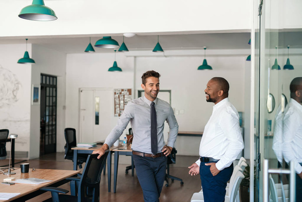 Dos jóvenes empresarios diversos hablando y riendo juntos mientras están de pie en una gran oficina moderna
 - Foto, Imagen