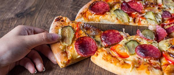 female hand takes a slice of Pepperoni Pizza with Mozzarella cheese, salami, bacon, Tomato sauce, pepper, Spices and pickled cucumbers. Italian pizza on wooden table background - Photo, Image