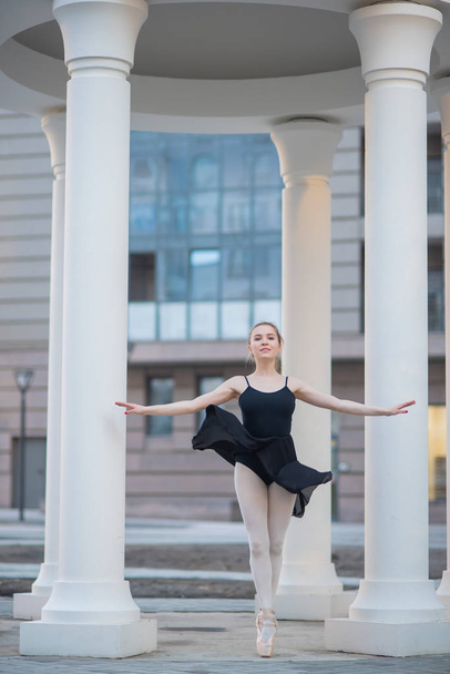 Ballerina in a black dress is a beautiful pose. A beautiful young woman in a black bodysuit is dancing in an elegant classical dance on a background of columns. - Fotografie, Obrázek