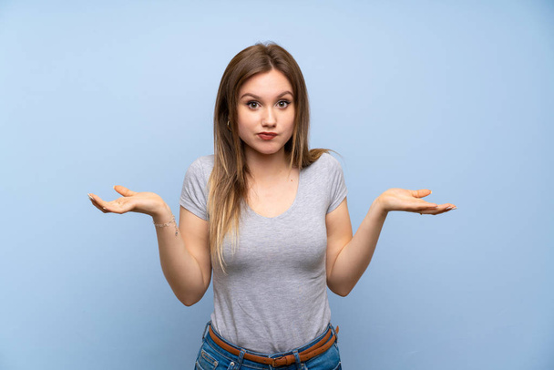 Teenager girl over isolated blue wall having doubts while raising hands - Фото, изображение
