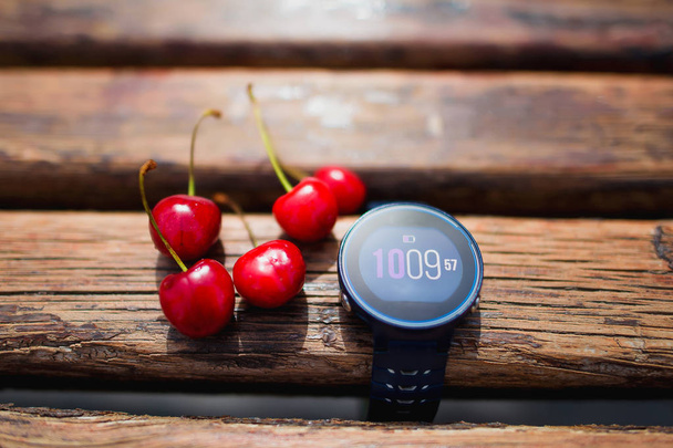 Fitness watches lie on a wooden bench. Red cherries. Hours for training. - Photo, Image