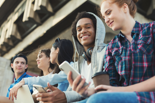 Πολυεθνική έφηβος φίλους που κάθονται σε εξωτερικούς χώρους, μιλούν και χρησιμοποιούν gadget - Φωτογραφία, εικόνα