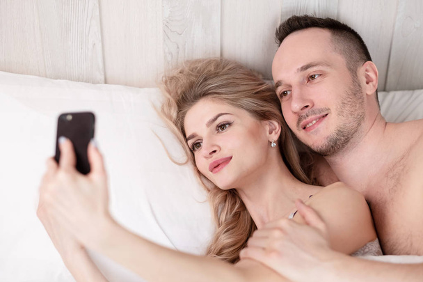 A married couple, a man and a woman, lie in bed, hug, embrace and take a selfie. - Fotografie, Obrázek