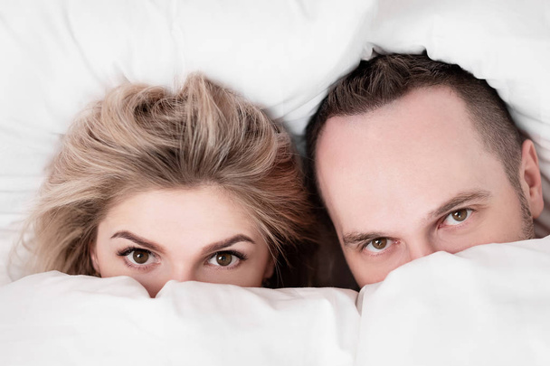 Married couple, man and woman, are lying in bed, hugging and sleeping on white bedding. Hiding their faces under blanket and looking out. - Photo, image