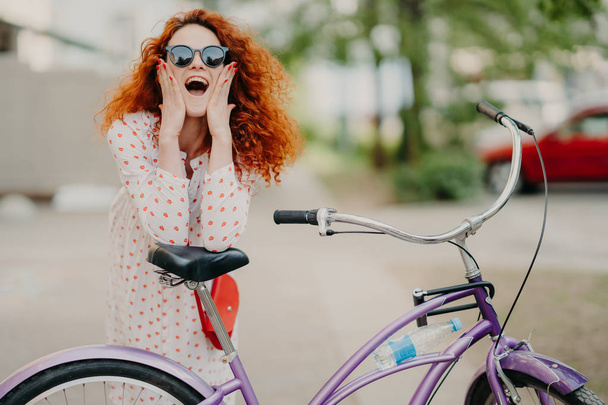 Cheerful female urban biker wears sunglasses and dress, has luxurious crisp foxy hair, keeps elbows on saddle of bicycle, spends recreation time in open air, poses over blurred street background - Photo, Image