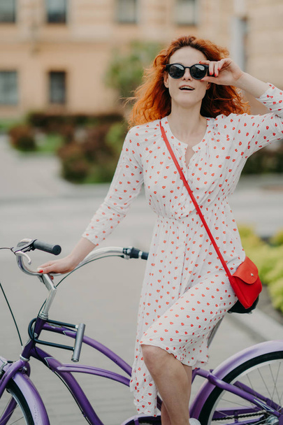 Styl uliczny i koncepcja aktywnego odpoczynku. Kręcone czerwone włosy kobieta trzyma rękę na ramie okulary, nosi białą sukienkę, koncentruje się na odległość, czeka na przyjaciela, będzie mieć na zewnątrz podróż na rowerze - Zdjęcie, obraz