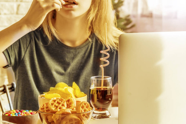 Το κορίτσι δουλεύει σε έναν υπολογιστή και τρώει φαστ φουντ. Ανθυγιεινά τρόφιμα: μάρκες, κράκερ, καραμέλες, βάφλες, κόκα κόλα. Πρόχειρο φαγητό, έννοια. - Φωτογραφία, εικόνα