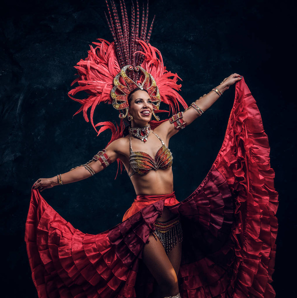 Ταλαντούχα χαρούμενη χορεύτρια με κόκκινο φτερό κοστούμι ποζάρει σε μικρό σκοτεινό στούντιο. - Φωτογραφία, εικόνα