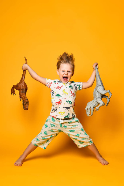 Küçük neşeli çocuk her iki elinde de plastik bir dinozor tutuyor. Çocuk üzerinde dinozor resimleri olan bir takım elbise giyiyor.. - Fotoğraf, Görsel
