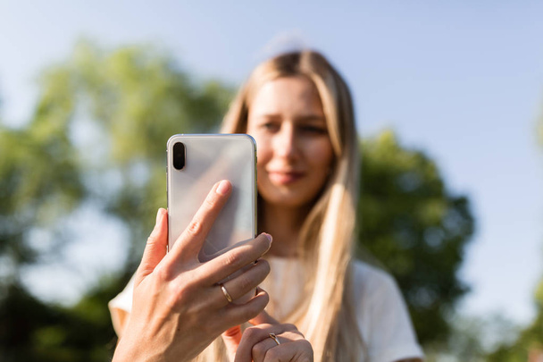 Hermosa mujer joven con cabello rubio utilizando el teléfono móvil al aire libre. Elegante chica haciendo selfie
 - Foto, Imagen