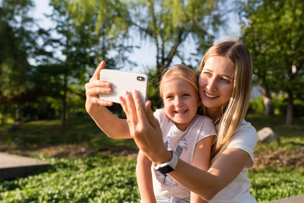 Молодые мама и дочь со светлыми волосами с помощью мобильного телефона на улице. Стильные девушки делают селфи в парке. Люди, использующие технологии
  - Фото, изображение