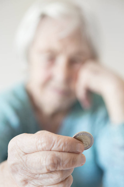 Triste anciana sentada a la mesa en casa y mirando miserablemente a solo la moneda restante de la pensión en su mano. - Foto, imagen