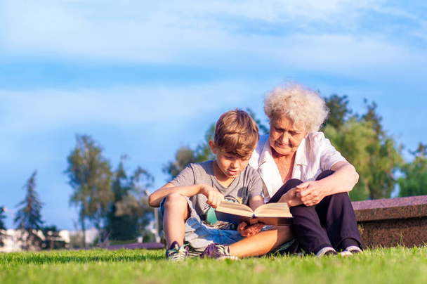 Привлекательная бабушка с внуком вместе читают книгу на улице. Летний семейный отдых. Самообразование и развитие
 - Фото, изображение