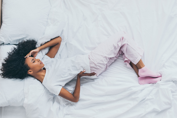 top view of unhappy african american woman suffering from back pain while lying on white bedding - 写真・画像