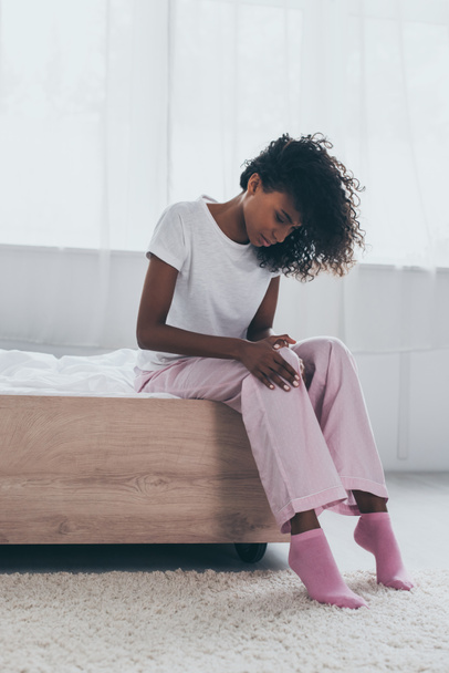 hübsche Afroamerikanerin leidet unter Schmerzen im Knie, während sie auf dem Bett sitzt  - Foto, Bild