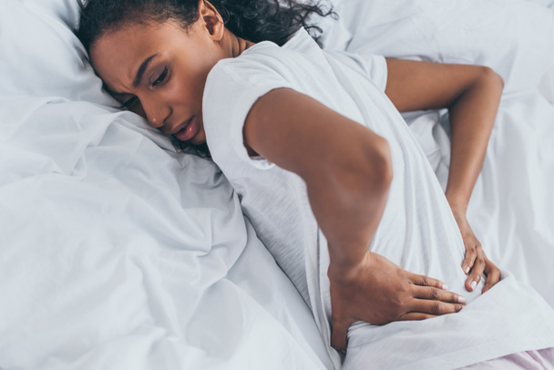 exhausted african american woman suffering from back pain while lying on bedding - Photo, image