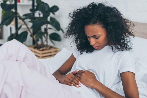 unhappy african american woman suffering from abdominal pain while lying in bed - Photo, Image
