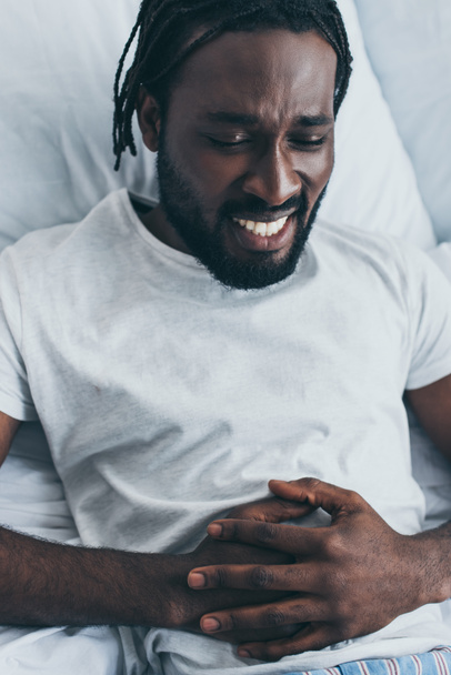 uitgeput Afro-Amerikaanse man lijdt aan maagpijn in de slaapkamer - Foto, afbeelding
