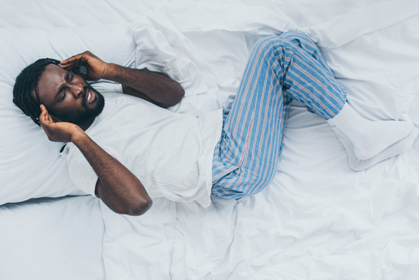top view of exhausted african american man suffering from headache while lying on white bedding - Фото, зображення