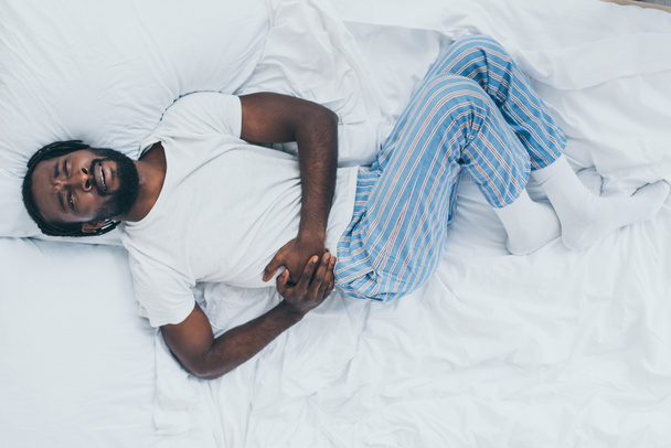 top view of exhausted african american man suffering from abdominal pain while lying in bed - 写真・画像