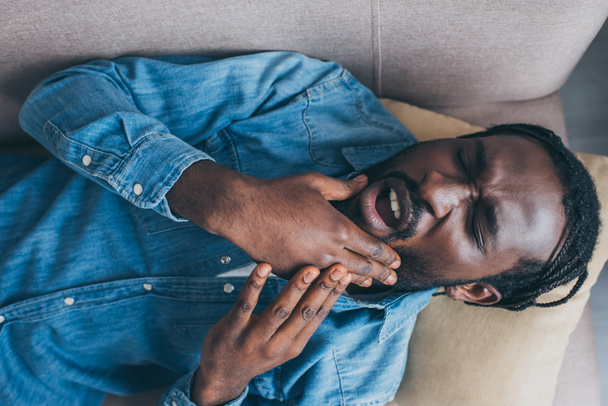 ソファに横たわり、顎の痛みに苦しむアフリカ系アメリカ人男性のトップビュー - 写真・画像
