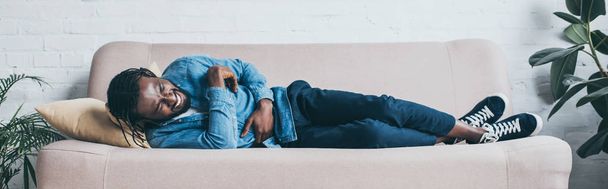 panoramic shot of african american man suffering from abdominal pain while lying on sofa - 写真・画像