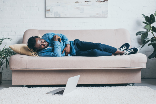 fiatal afro-amerikai férfi szenved gyomorfájdalom, miközben fekszik a kanapén közelében laptop padlón - Fotó, kép