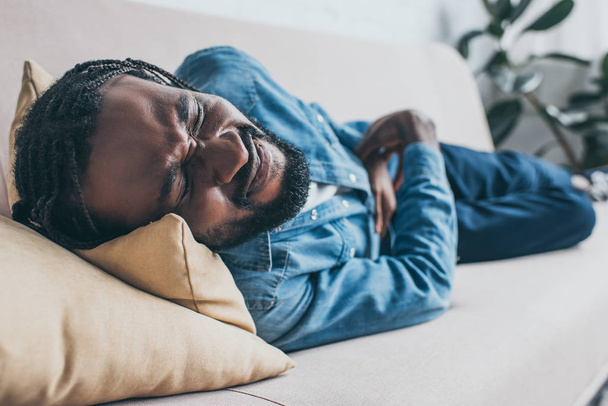 selective focus of exhausted african american man lying on sofa and suffering from abdominal pain - 写真・画像