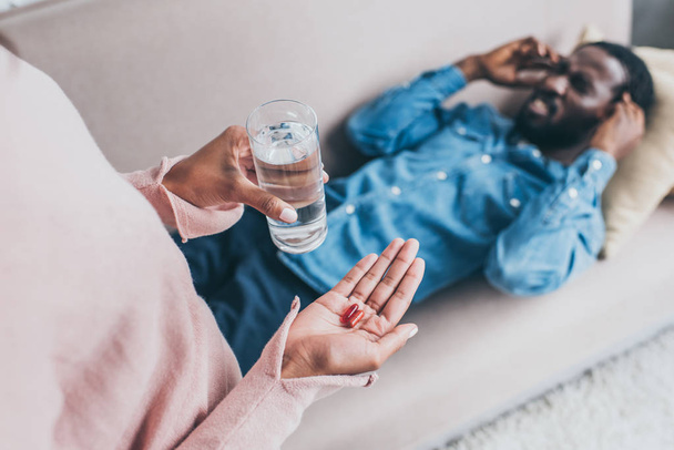 cropped view of african american woman holding glass of water and pills while standing near man suffering from headache on sofa - Fotografie, Obrázek