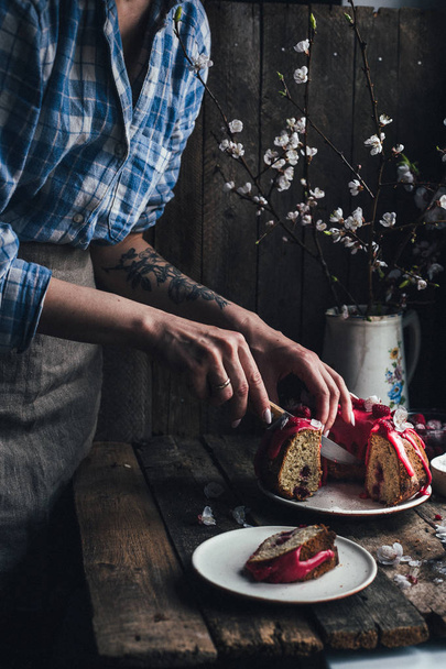 木製のテーブルの上に白い花とラズベリーバンドケーキを切る女性のトリミングビュー, 選択的な焦点 - 写真・画像