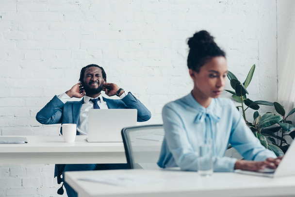 hombre de negocios afroamericano que sufre de dolor de cabeza mientras está sentado en la oficina cerca de gerente bastante
 - Foto, Imagen