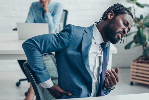 unglücklicher afrikanisch-amerikanischer Geschäftsmann leidet unter Rückenschmerzen, während er in der Nähe seines Kollegen im Büro sitzt - Foto, Bild