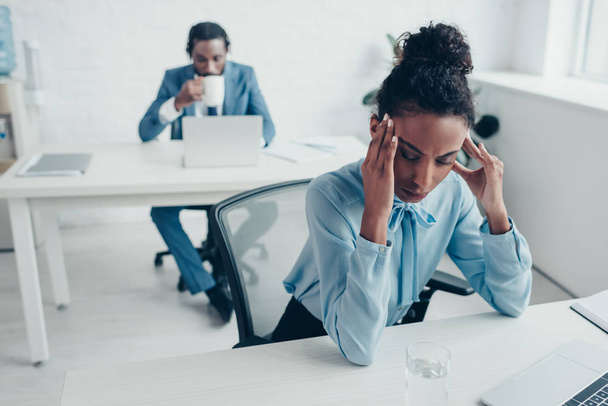 enfoque selectivo de la mujer de negocios afroamericana que sufre de dolor de cabeza mientras está sentada en la oficina cerca de colega beber café
 - Foto, Imagen