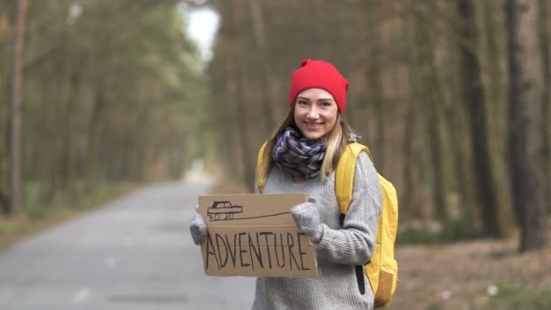 Młoda ładna autostopowiczka tańczy na drodze z plakatem Adventure .Travel emocji - Materiał filmowy, wideo