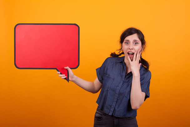 Shocked young woman with a speech bubble against a yellow background in studio - Photo, Image