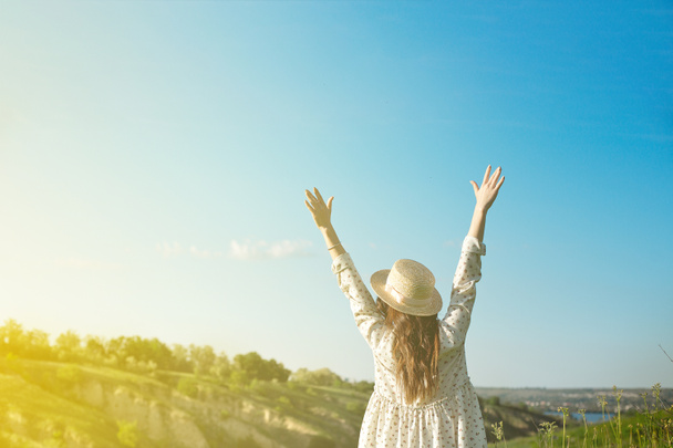 Una donna felice con un cappello di paglia a braccia aperte rimane sul bordo di una scogliera sotto la luce del tramonto del cielo, godendo di successo, libertà e un futuro luminoso
 - Foto, immagini