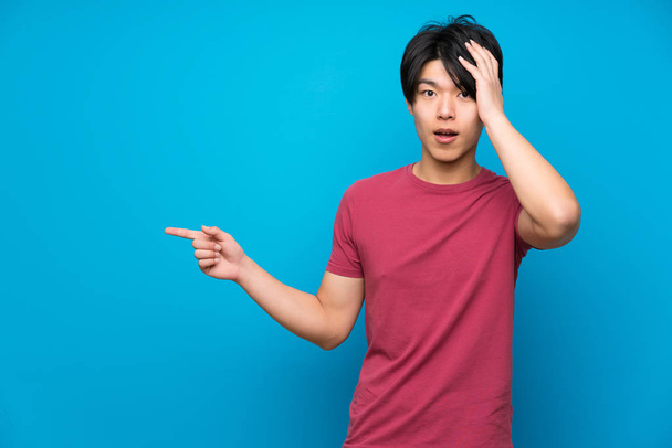 Asiatique homme avec chemise rouge sur isolé mur bleu pointant doigt sur le côté et présentant un produit
 - Photo, image