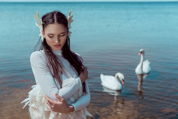 tender woman in white swan costume closing eyes, standing near river with birds - Photo, Image