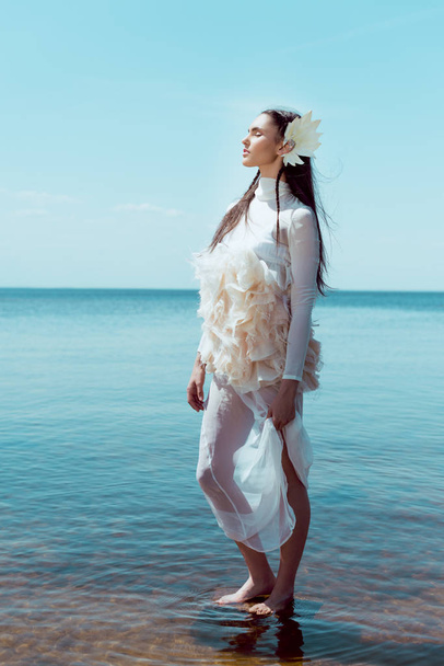 young woman in white swan costume standing on river and sky background, closing eyes - Photo, Image