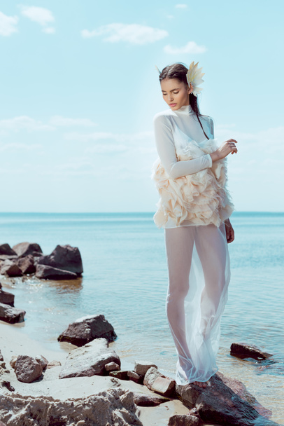 Tender vrouw in witte zwaan kostuum op zoek weg, staande op het strand in de buurt van water - Foto, afbeelding