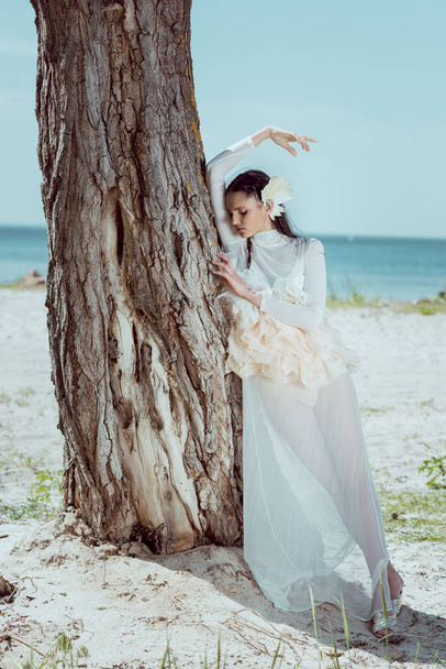 Beyaz kuğu kostümlü genç bir kadın sahilde üç sandığın yanında duruyor. - Fotoğraf, Görsel