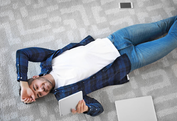 Young man with modern devices lying on soft carpet at home - Fotoğraf, Görsel