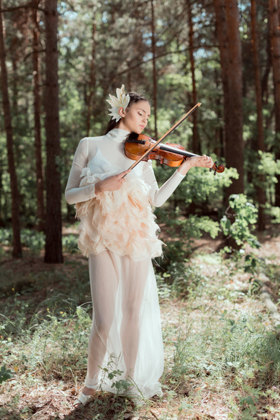 elegante vrouw in witte zwaan kostuum staande op forest achtergrond, spelen op viool - Foto, afbeelding